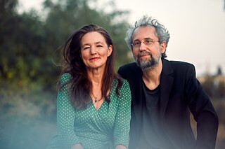 Die beiden Podcasthosts: Links im Bild Judith Gridl im grünen, weiß-gepunkteten Kleid. Rechts neben ihr Klaus Rathje mit schwarzen Sakko, grauen Bart und grauen Haaren.