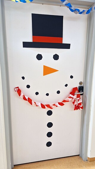 An einer weißen Innentüre hängen Girlanden, ein Zilinder und ein stilisiertes Gesicht.