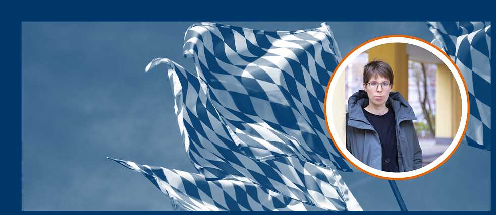 Im Hintergrund sieht man die Fahnen Bayerns im Wind wehen. Im Vordergrund ist ein Bild mit der Autorin Bettina Wilpert. Sie ist weiß, trägt kurze braune Haare, eine dicke Jacke über einem Shirt und Brille.