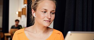 Tatjana in einer Lernsituation. Sie sitzt in einem Café, trägt Kopfhörer und blickt auf ein Tablet.