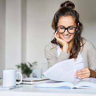 Eine lächelnde Frau sitzt an einem Schreibtisch und liest in einem Lehrbuch.