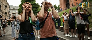 Gruppe von Personen, zwei davon im Vordergrund, spazieren in einem urbanen Umfeld mit Kopfhörern und Fingern um die Augen wie eine Brille