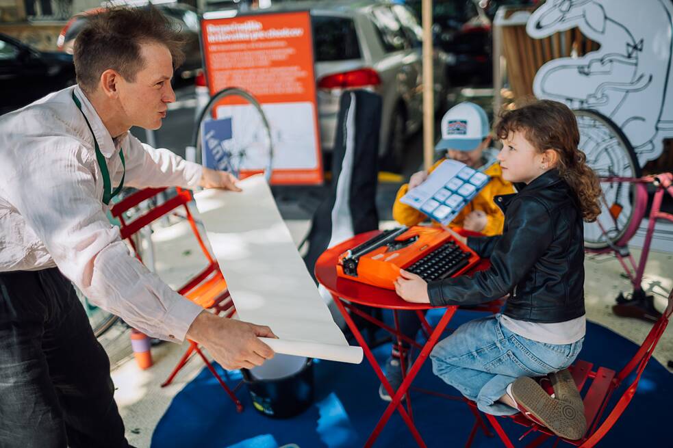 Artist with a girl at a type writer handing her a roll of paper