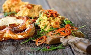 A pretzel lies on a wooden table. Next to it on a long wooden board are three scoops of yellow cheese spread.