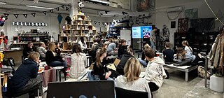 A large workshop space with high ceilings and industrial lighting, where a group of people are seated and attentively watching a presentation.