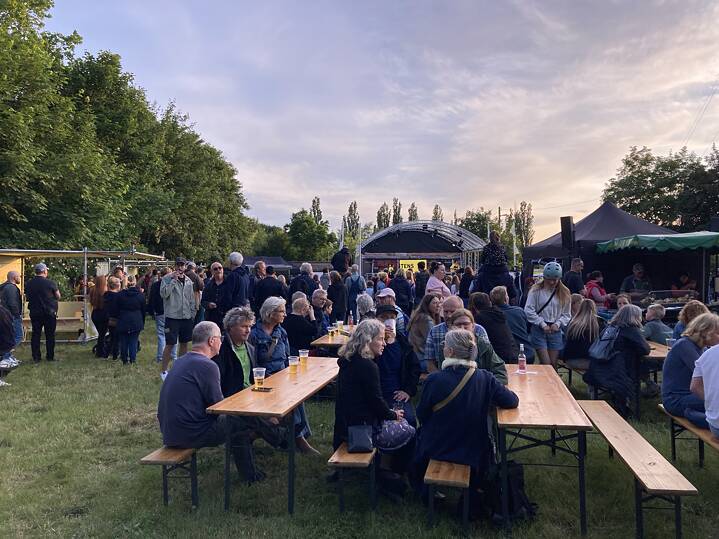 Menschen sitzen bei Sonnenuntergang auf Holzbänken und -tischen im Freien, umgeben von Bäumen, im Rahmen des Garagen-Festivals