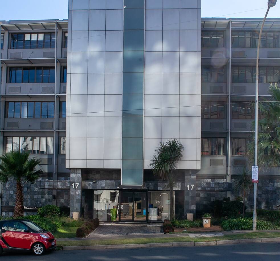 Das Bild zeigt die Vorderansicht eines modernen Gebäudes mit einer Fassade aus Glas und Metall. Das Gebäude hat mehrere Etagen mit großen Fenstern. Links und rechts neben dem Eingang stehen jeweils eine Palme. Die Zahl „17“ ist beidseitig neben dem Eingang gut sichtbar angebracht.