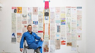 Ein Mann, der vor einer Wand sitzt, die dicht mit verschiedenen Papieren bedeckt ist, die ausgestellt werden sollen.