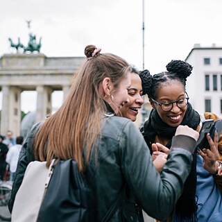 Drei junge Frauen schauen lachend auf einen Telefonbildschirm, im Hintergrund ist das Brandenburger Tor in Berlin zu sehen.