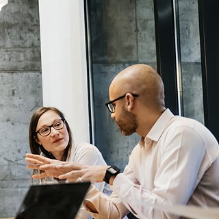 Junge Frauen und Männer sitzen zu einer Besprechung an einem Tisch. Ein Mann spricht und gestikuliert.