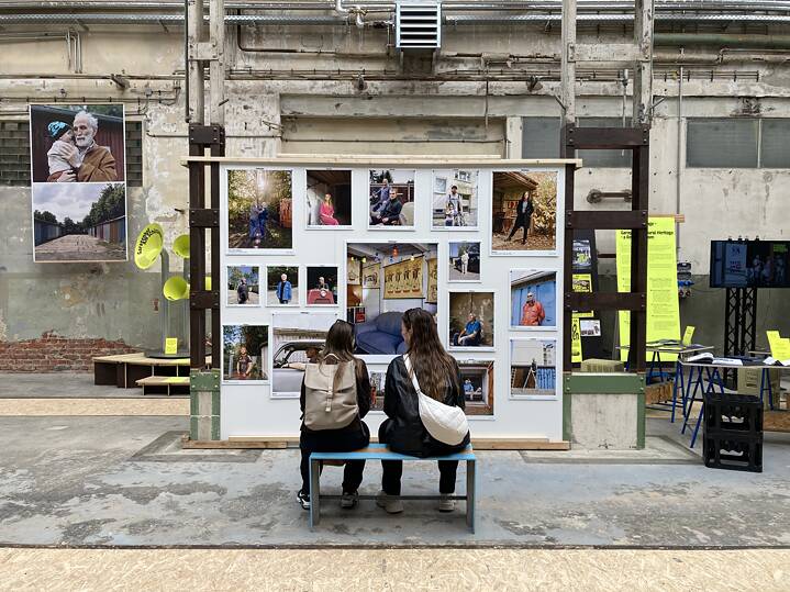 Im Vordergrund: Zwei Personen sitzen auf einer Bank und schauen sich Fotos der Ausstellung #3000Garagen an, die im Garagen-Campus hängen