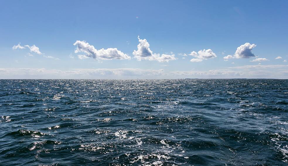 A wide view of the open ocean under a clear blue sky, with small waves . Fluffy white clouds are scattered across the distant sky, and the horizon line cleanly separates sea and sky