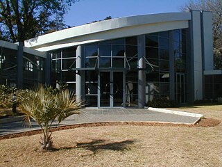 Das Bild zeigt die Außenansicht des Neubaus vom  Goethe-Instituts Johannesburg. Die Fassade besteht aus großen Glasfenstern, und das Dach ist elegant geschwungen. Der Eingangsbereich verfügt über doppelte Glastüren, die von zwei senkrechten Säulen eingerahmt und gestützt werden.  Vor dem Gebäude befindet sich eine kleine, gepflegte Grünfläche mit Rasen und einer palmenähnlichen Pflanze. Die Umgebung wirkt ordentlich und einladend.