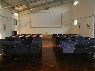 Das Bild zeigt das leere Auditorium. Mehrere Reihen blauer Stühle sind auf den vorderen Bereich des Raumes ausgerichtet, wo sich eine große weiße Projektionsfläche befindet. Links neben der Leinwand steht ein Rednerpult.  Der Raum hat eine hohe Decke mit sichtbaren Metallträgern. An den Wänden hängen gerahmte Bilder, und mehrere kleine Fenster lassen Tageslicht herein.