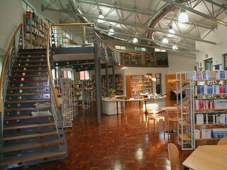 Das Bild zeigt den Innenraum des Infocenters. Auf der linken Seite befindet sich eine große, geschwungene Treppe, die zu einer oberen Etage mit weiteren Bücherregalen führt.  Im Erdgeschoss stehen mehrere Tische und Stühle, die zum Lesen oder Arbeiten genutzt werden können. Die Umgebung ist von zahlreichen Bücherregalen umgeben, die mit Büchern gefüllt sind.  Die Decke ist offen gestaltet mit sichtbaren Metallträgern. Hängende Leuchten sorgen für eine gleichmäßige und helle Ausleuchtung des Raumes.