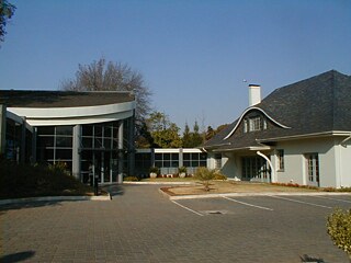 Das Bild zeigt einen modernen Gebäudekomplex, bestehend aus zwei Gebäuden, vor denen gepflasterter Innenhof liegt. Sie sind durch einen verglasten Gang verbunden.  Das linke Gebäude hat eine moderne Glasfassade mit großen Fenstern und ein geschwungenes Dach. Das rechte Gebäude wirkt traditioneller, ebenfalls mit einem geschwungenen Dach und mehreren rechteckigen Fenstern.  Im Vordergrund des Bildes befindet sich ein Parkplatz mit klar markierten Stellplätzen. Im Hintergrund umgeben Bäume und Sträucher den Gebäudekomplex und schaffen eine grüne, gepflegte Umgebung.