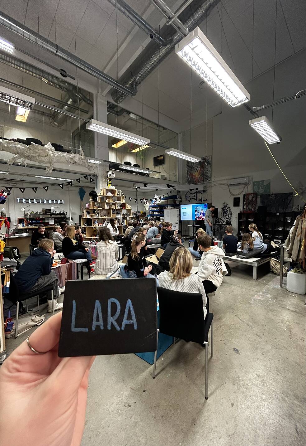A large workshop space with high ceilings and industrial lighting, where a group of people are seated and attentively watching a presentation. In the foreground, a hand holds a small blackboard sign with the name LARA written in blue chalk