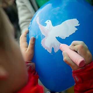 Ein Junge malt eine weiße Taube auf einen blauen Luftballon. 