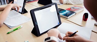 Group of students writing in notebooks and using tablets at a classroom table. © ©Goethe-Institut Deutsch 2.0
