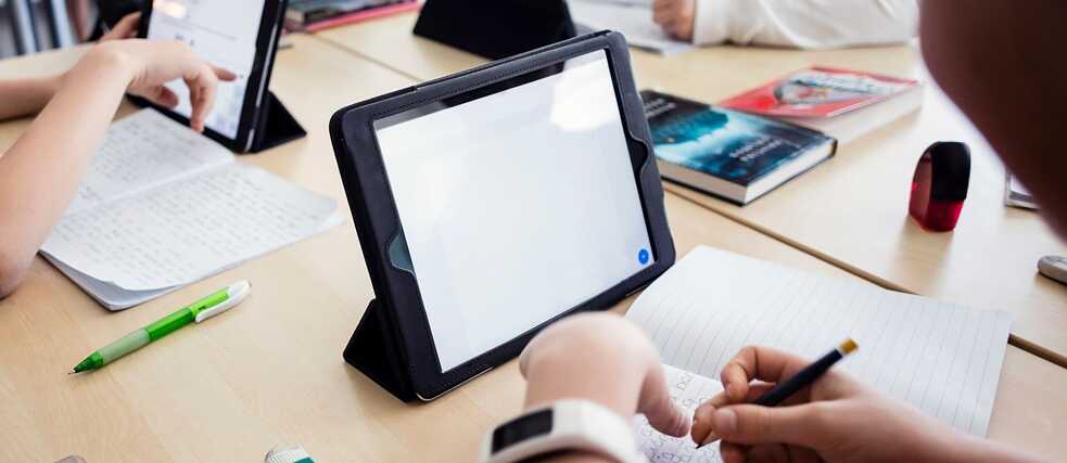 Group of students writing in notebooks and using tablets at a classroom table.