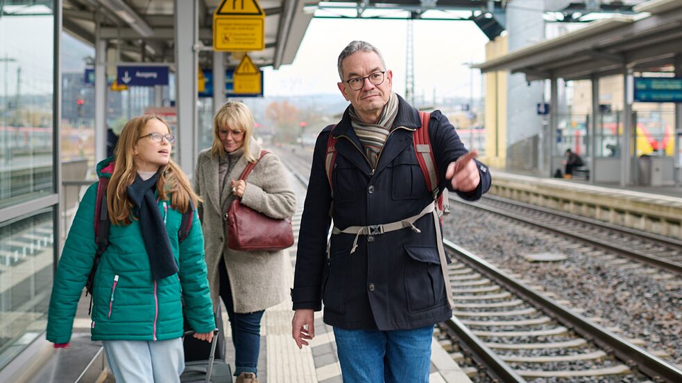 Eine Familie geht auf einem Bahnsteig entlang, im Hintergrund steht ein Zug.