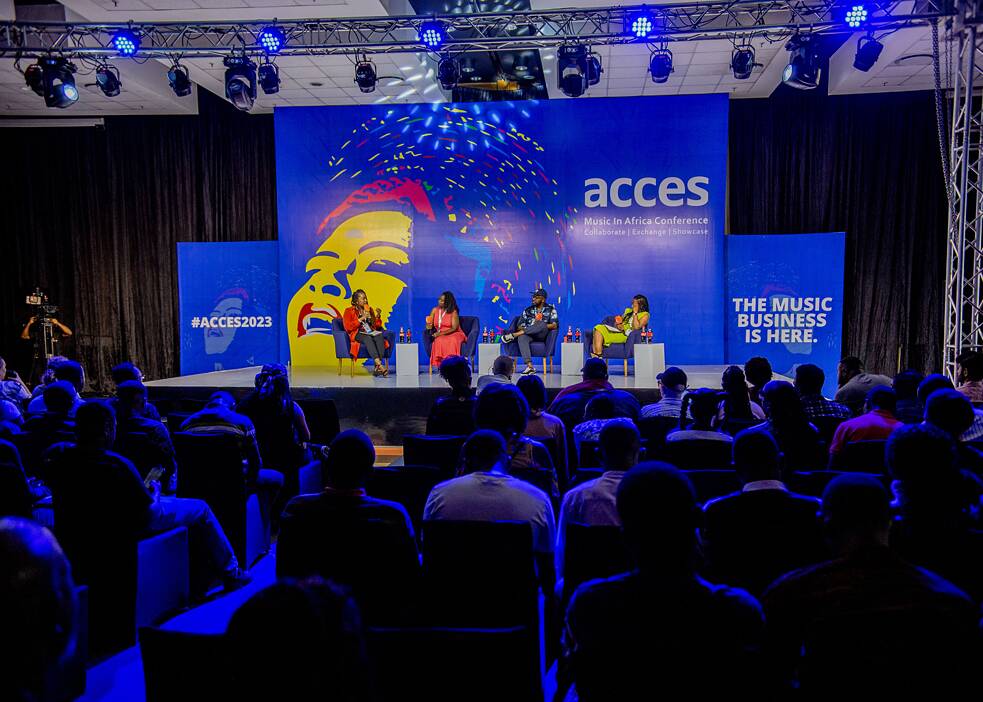 Das Bild zeigt eine Podiumsdiskussion im Rahmen der Veranstaltung ACCES – Music in Africa Conference for Collaborations, Exchange and Showcases. Auf der Bühne sitzen vier Podiumsteilnehmende. Hinter ihnen befindet sich ein großes, farbenfrohes Bühnenbanner mit einer künstlerischen Darstellung eines Gesichts sowie dem Schriftzug „acces“ und dem Hashtag „#ACCES2023“.  Rechts auf dem Banner steht in großen Buchstaben: „The Music Business is here“. Im Vordergrund ist das Publikum zu sehen, das der Diskussion zugewandt ist.