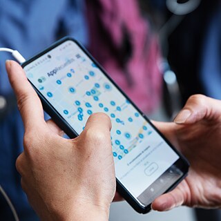 Primer plano de unas manos sosteniendo un teléfono inteligente. En la pantalla se muestra un mapa con numerosos marcadores azules. En la parte superior aparece la palabra "App" seguida de texto parcialmente oculto. En la parte inferior hay un campo de entrada o botón. Un cable blanco está conectado en la esquina superior izquierda del dispositivo.