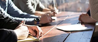 A round table, visible are hands, somebody writing, a note-pad on the table