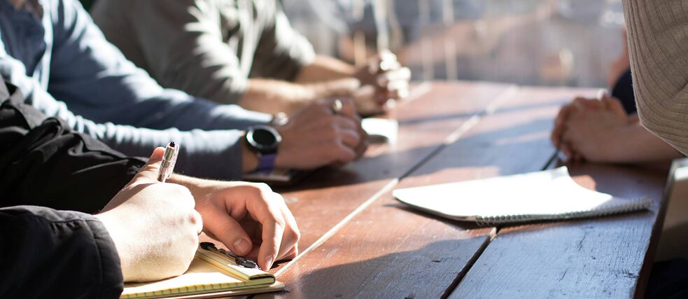A round table, visible are hands, somebody writing, a note-pad on the table