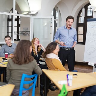 Mehrere Teilnehmende sitzen gemeinsam in einem modernen Seminarraum. Eine Lehrkraft steht am Whiteboard und erklärt Inhalte anhand einer Mindmap. Die Atmosphäre ist offen, konzentriert und interaktiv.