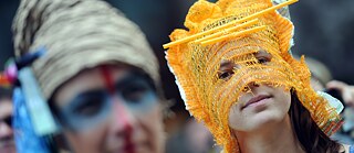 Zwei junge Frauen demonstrieren bei der Fashion Week Berlin für soziale, ökologische und faire Kleidung. Sie tragen phantasievolle, selbst gestaltete Kopfbedeckungen aus Gemüse-Netzen.