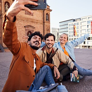 Eine Gruppe von drei Personen posiert fröhlich für ein Selfie vor einem beeindruckenden Gebäude