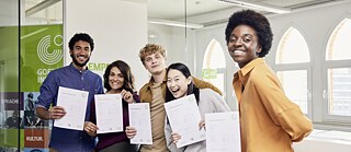 Eine bunt gemischte Gruppe junger Leute, die freudig Goethe-Zertifikate hochhält, lächelt und miteinander ins Gespräch kommt