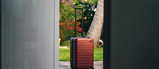 A red hardshell suitcase with wheels stands on a concrete surface just outside an open door, framed by a lush green garden with palm trees and vibrant flowers in the background. &copy; © Unsplash A Red Suitcase Ready for Travel Outside a Door