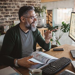 Ein Mann sitzt am Schreibtisch und hat eine Tastatur vor sich. Mit einem Bleistift in der Hand zeigt er auf seinen Bildschirm.