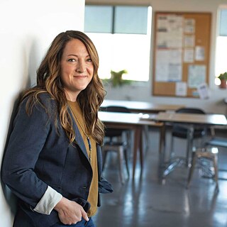 Eine Lehrerin lehnt an einem offenen Türrahmen. Im Hintergrund sieht man leicht verschwommen ein Klassenzimmer