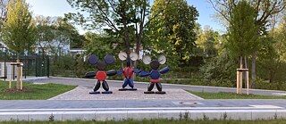 Drei skulpturale Figuren aus runden und zylindrischen Metallteilen stehen in einer Reihe auf einem gepflasterten Platz im Freien. Die mittlere Figur trägt ein rotes Oberteil und blaue Hosen, während die äußeren Figuren schwarze Beine haben