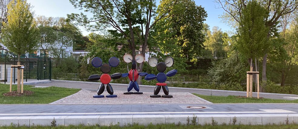 Drei skulpturale Figuren aus runden und zylindrischen Metallteilen stehen in einer Reihe auf einem gepflasterten Platz im Freien. Die mittlere Figur trägt ein rotes Oberteil und blaue Hosen, während die äußeren Figuren schwarze Beine haben