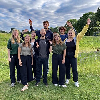 Eine kleine Gruppe junger Menschen steht auf einer Wiese und grüßt in die Kamera.