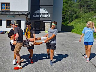 Jugendliche z.T. mit verbundenen Augen tragen einen jungen Mann über einen Hof.