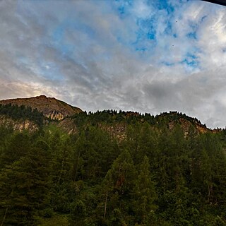 Blick auf einen bewalteten Berg, im Hintergrund sich aufläsende Gewitterwolken