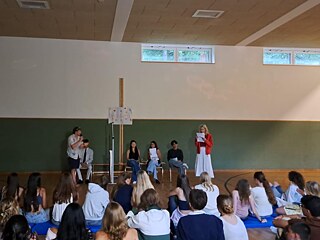 Jugendliche auf einer in einer Turnhalle improvisierten Bühne, zwei haben ein Mikrophon in der Hand.