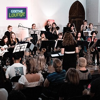 This image shows a vibrant orchestral performance in an intimate indoor venue. A group of young female musicians plays in a semi-circle, led by an engaged female conductor. The close audience and soft lighting create a warm, communal feel. The setting highlights tradition, mentorship, and the shared joy of live music.