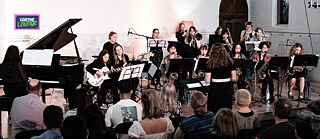 This image shows a vibrant orchestral performance in an intimate indoor venue. A group of young female musicians plays in a semi-circle, led by an engaged female conductor. The close audience and soft lighting create a warm, communal feel. The setting highlights tradition, mentorship, and the shared joy of live music.