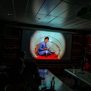 A projection screen lights up a dark room with people looking at it around a desk. The projection slide shows someone sat in a tent reading a book