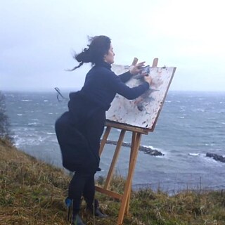 A woman painting at an easel leaning towards the edge of a cliff with strong wind blowing her hair back