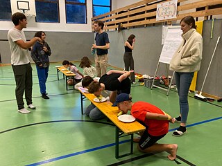 Jugendliche in einer Turnhalle kniehen vor einer Bierbank und versuchen, eine Banane zu essen, mit verbundenen Handen auf dem Rücken.