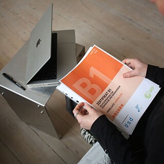 Eine Deutschlehrerin sitzt vor einem Computer und hält ein Goethe-Zertifikat B1 in den Händen.