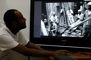 Eine Person sitzt an einem Schreibtisch und blickt auf einen großen Monitor. Auf dem Bildschirm ist ein schwarz-weißes Foto zu sehen, das mehrere Menschen bei der Arbeit mit Metallstangen und anderen Materialien in einem industriellen oder baulichen Umfeld zeigt. Die Person am Schreibtisch bedient einen Computer mit Maus und Tastatur. 
