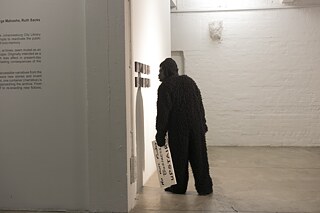 In einem Ausstellungsraum steht eine Person in einem Gorilla-Kostüm. Sie hält ein Schild mit einer Aufschrift in der Hand, die nicht gut zu lesen ist. Die Person blickt auf mehrere kleine, gerahmte Kunstwerke, die an der Wand hängen.